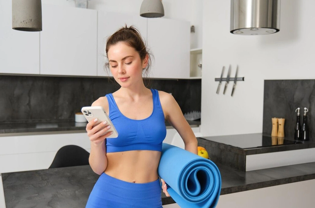 "Woman in blue workout gear checking phone with a yoga mat, showcasing activewear fabric quality and comfort, ideal for sportswear made from GSM fabrics."