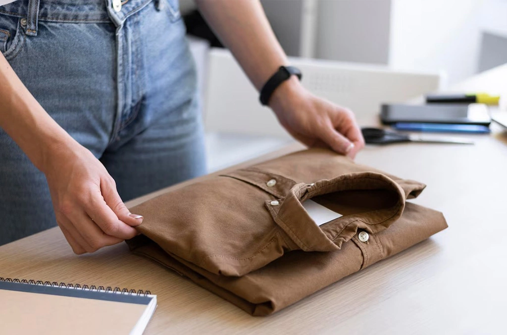 "Person folding brown shirt, emphasizing the practicality and texture of fabrics with specific GSM for comfort and performance in sportswear."